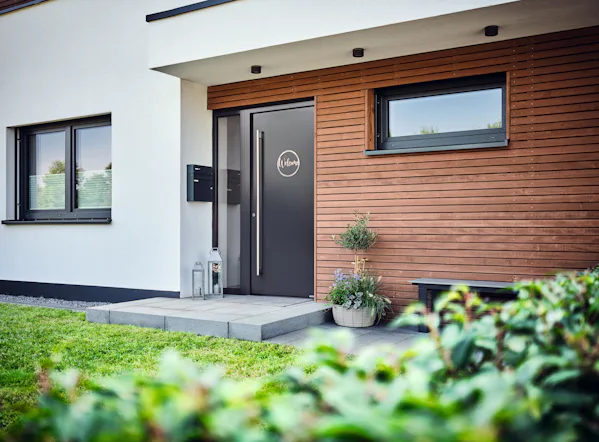 Op de afbeelding is de moderne entree van een woning in Friesland te zien, voorzien van een strakke antracietkleurige voordeur van het merk Hörmann. Op de deur staat in een rond raam het woord "Welcome" geschreven. De gevel van de woning is deels wit gestuct en deels bekleed met horizontale houten latten. Naast de deur staat een plantenpot met een sierlijke plant en een decoratief lantaarn. Links van de voordeur hangen een zwarte brievenbus en een deurbelpaneel. De woning straalt een nette en eigentijdse stijl uit, waarbij kwaliteit en design centraal staan – passend bij het imago van Hörmann, leverancier van hoogwaardige garagedeuren en voordeuren.