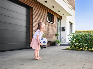 Ruimtebesparing Kind in roze rok speelt met voetbal voor een LPU 42 garagedeur sectionaal van Hörmann. Het huis is met hout afgewerkt en rechts ziet u groene struiken.