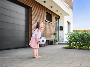 Ruimtebesparing Kind in roze rok speelt met voetbal voor een LPU 42 garagedeur sectionaal van Hörmann. Het huis is met hout afgewerkt en rechts ziet u groene struiken.