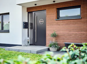 ThermoSafe voordeur met kozijn | Hörmann Zwarte ThermoSafe voordeur met kozijn in modern huis met houtaccenten, op de voorgrond is gras en een struik te zien