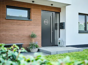 ThermoSafe voordeur met kozijn | Hörmann Zwarte ThermoSafe voordeur met kozijn in modern huis met houtaccenten, op de voorgrond is gras en een struik te zien