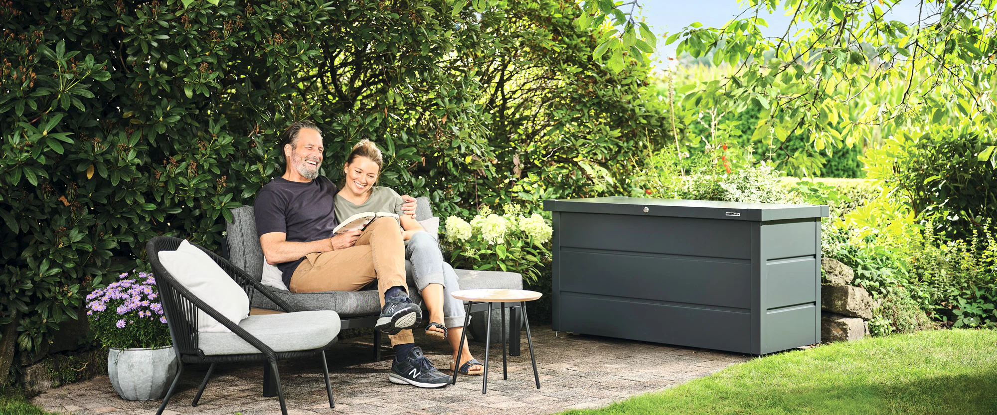 Een man en een vrouw zitten op een loungestoel is een groene tuin en naast hun staat een kussenbox.