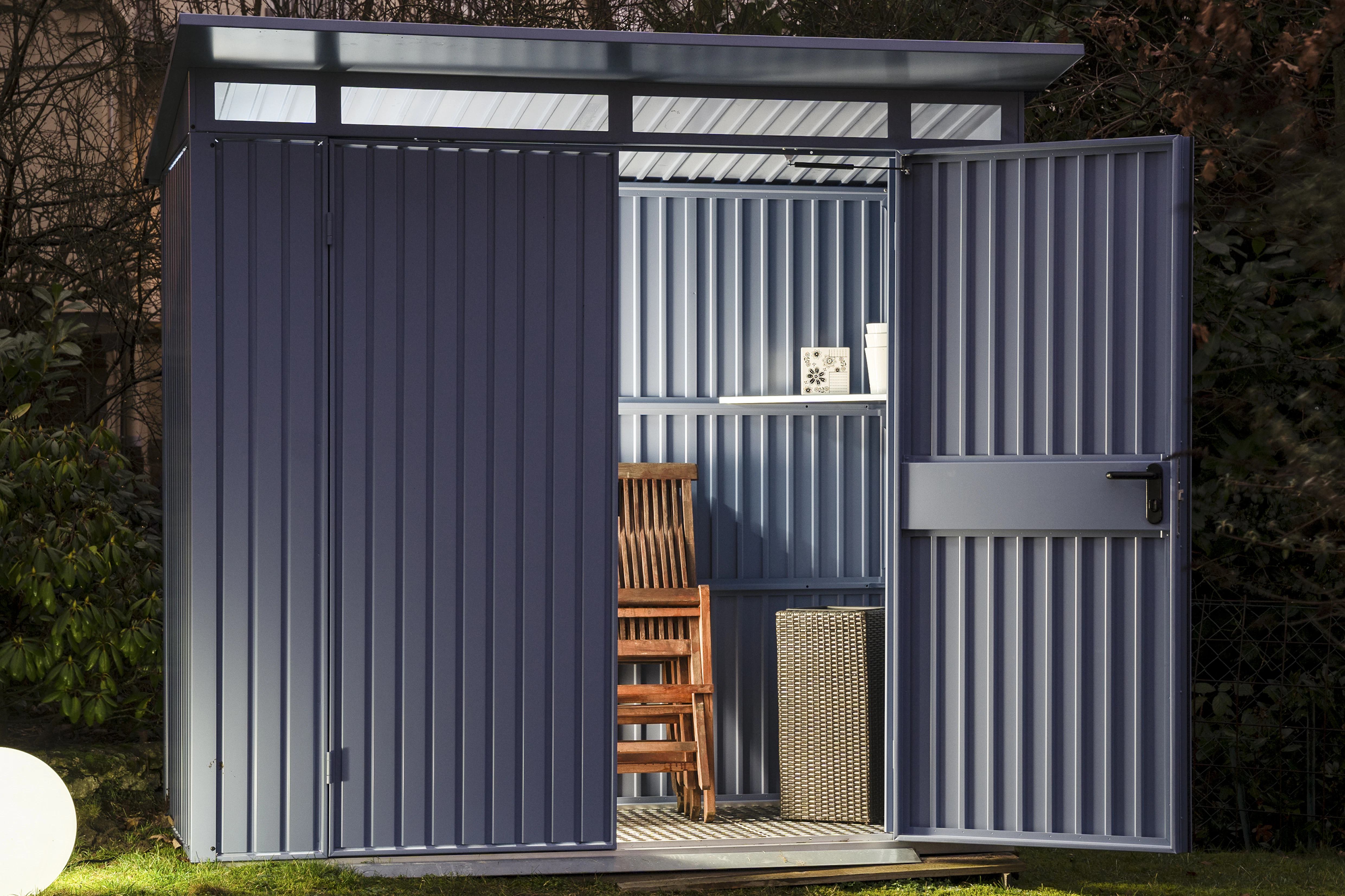 Blauwkleurig tuinhuis van Hörmann verlicht door een ronde lamp. Binnen in het tuinhuis staan spullen opgeborgen waaronder een houten tuinstoel.