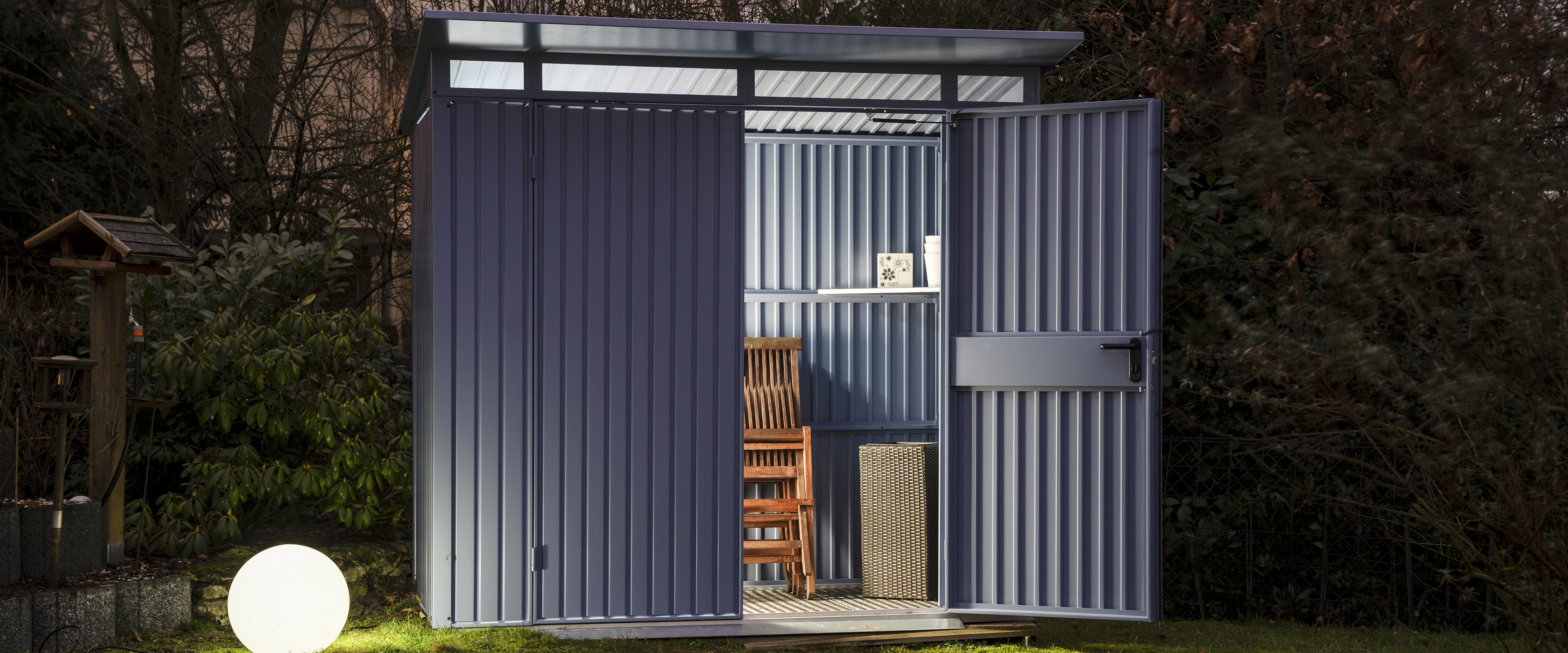 Blauwkleurig tuinhuis van Hörmann verlicht door een ronde lamp. Binnen in het tuinhuis staan spullen opgeborgen waaronder een houten tuinstoel.