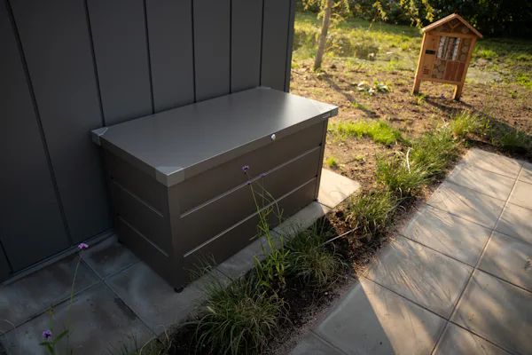 Juno Waterdichte Kussenbox op terras | Hörmann Waterdichte kussenbox met metalen deksel naast een moderne gevel. Op de achtergrond een insectenhotel in een groene tuinomgeving.