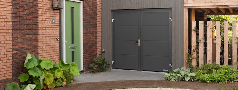 Antraciet openslaande garagedeur van Hörmann met een houten stalling ernaast, met een groene voordeur en beplanting naast het huis.