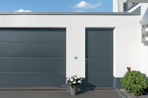 Tips voor het kiezen van de juiste garagedeur met loopdeur Strakke witte gevel met grijze Hörmann garagedeur en bijpassende loopdeur, bloempot en huisnummer 6 naast de deur.