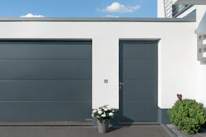 Tips voor het kiezen van de juiste garagedeur met loopdeur Strakke witte gevel met grijze Hörmann garagedeur en bijpassende loopdeur, bloempot en huisnummer 6 naast de deur.