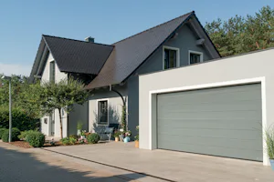 De voordelen van een elektrische garagedeur voor nieuwbouwwoningen Grijze woning met zwarte kap en grote grijze Hörmann sectionaaldeur, omringd door planten en netjes aangelegde oprit.