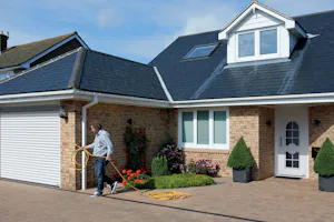 Tips voor het kiezen van de juiste roldeur voor uw werk- of hobbyruimte Bakstenen woning met witte Hörmann roldeur, man met tuinslang op oprit en nette voortuin met bloemen.