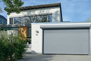 Belangrijke veiligheidskenmerken van Strakke witte woning met gesloten lichtgrijze Hörmann roldeur en betegelde oprit, omgeven door groen.