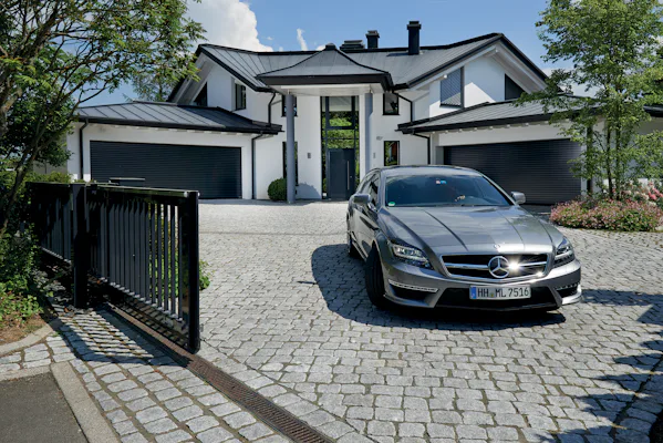Een auto rijdt een open hek uit. Achter ziet u een luxe villa met twee garages, in de garages zitten RollMatic roldeuren van Hörmann.