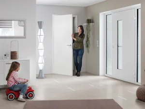 Voordelen van een dubbele voordeur Witte dubbele Hörmann voordeur met glasstroken in moderne hal, vrouw bij binnendeur en kind op rood speelgoedautootje.