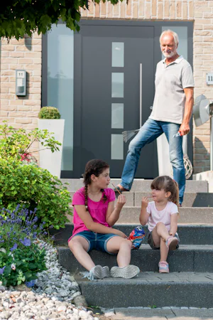 ThermoSafe voordeur | Hörmann Gezellige gezinssfeer voor een moderne woning met een stijlvolle zwarte voordeur. Twee jonge meisjes zitten lachend op de traptreden, terwijl een oudere man ontspannen op de achtergrond staat. De voortuin is prachtig aangelegd met kleurrijke bloemen en groen, wat de woning een uitnodigende uitstraling geeft. Deze voordeur draagt bij aan de verhoging van de woningwaarde.