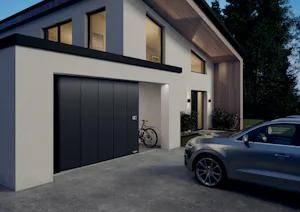 Technologische innovaties in elektrische garagedeuren die bijdragen aan comfort Moderne woning met zwarte zijdelingse Hörmann sectionaaldeur, geparkeerde fiets en zilveren auto op de oprit.
