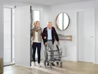 Nooit meer zoeken naar sleutels - open gewoon uw voordeur... Een jonge vrouw helpt een oudere man met een rollator bij het betreden van een woning via een brede, drempelloze deur.