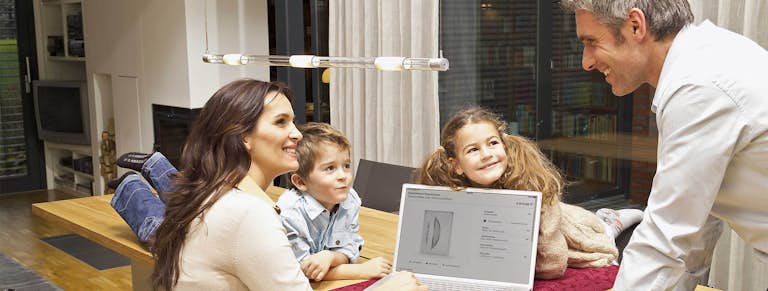 En familj sitter och kollar på datorn vid köksbordet.