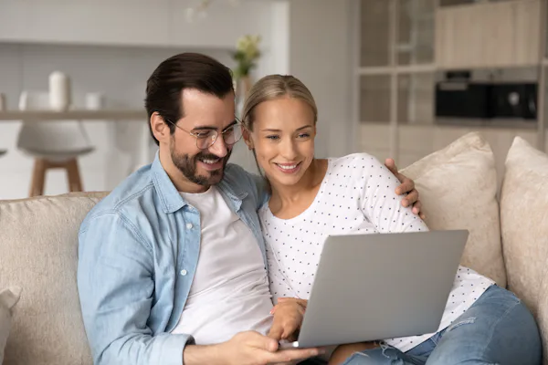 Een man en een vrouw zitten samen op een beige bank en kijken naar een laptop.