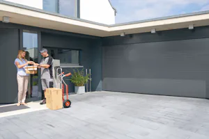 Enkele voordelen van onze Een vrouw ontvangt pakketjes bij een Hörmann ThermoCarbon voordeur naast een moderne garage en grijze gevel.