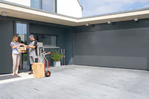 Enkele voordelen van onze Een vrouw ontvangt pakketjes bij een Hörmann ThermoCarbon voordeur naast een moderne garage en grijze gevel.