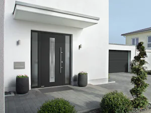 Enkele voordelen van de Een antracietkleurige voordeur Thermo65 in een wit modern huis. De voordeur heeft een raam midden in de deur en zijlichten. Verder ziet u begroeiing, een oprit en een garage.