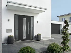 Enkele voordelen van de Een antracietkleurige voordeur Thermo65 in een wit modern huis. De voordeur heeft een raam midden in de deur en zijlichten. Verder ziet u begroeiing, een oprit en een garage.