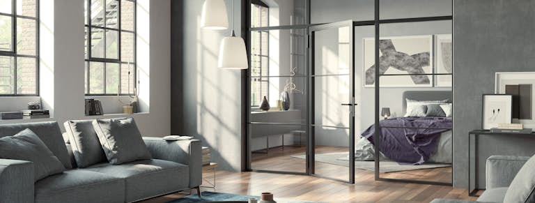 Das Bild zeigt eine modern eingerichtete Loft-Wohnung mit hohen Decken und einer Stahl-Glas-Trennwand mit Glastür. Genannt wird der Wohn- und Einrichtungsstil dieses Apartments „Industrial Style“.