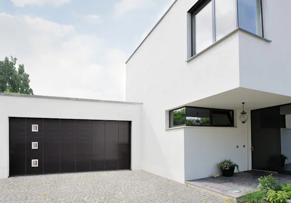 Prelis L Moderne witte woning met zwarte Hörmann garagedeur met loopdeur en vierkante glaspanelen op een stenen oprit.