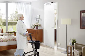 Elektrische deuropener | Ideaal voor ouderen Elektrische deuropener - Een oudere vrouw met een rollator loopt door een openstaande witte deur in een sfeervolle slaapkamer. De deur wordt automatisch geopend door een elektrische deuropener aan de bovenzijde van de deur. De kamer heeft een warme inrichting met een houten dressoir, decoratieve foto's aan de muur, een staande lamp en een comfortabele stoel.