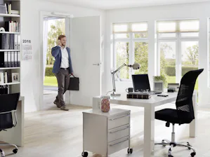 Elektrische deuropener | Toegepast in een kantoor Elektrische deuropener - Een man in zakelijke kleding loopt een modern, licht kantoor binnen terwijl hij zijn telefoon gebruikt. De deur wordt automatisch geopend door een elektrische deuropener. Het kantoor heeft een strak design met een witte bureautafel, zwarte bureaustoelen, een ladekast en grote ramen met uitzicht op een groene tuin.