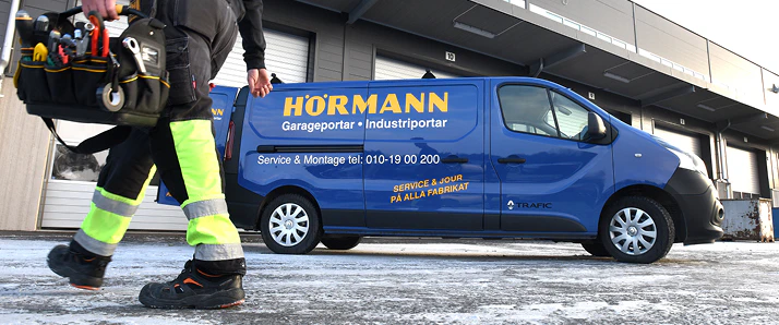 Servicetekniker på jobb. Går framför en blå Hörmann-servicebuss. i Bakgrunden är en industrifastighet med portar.