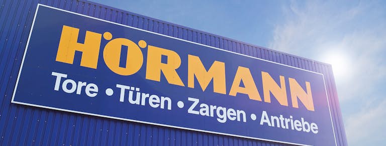 Het logo van Hörmann op een blauw pand met een zonnige lucht op de achtergrond.