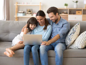 Ihre Vorteile im Überblick Vater, Mutter und Tochter sitzen auf einer Couch und schauen lächelnd in den Laptop.