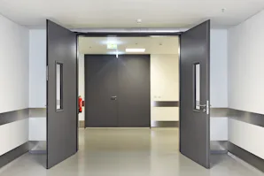 Dubbele binnendeuren met glas Dubbele grijze Hörmann binnendeuren met glazen kijkvensters, geopend naar een lichte gang in een moderne bedrijfsomgeving, voorzien van brandveiligheidssystemen en hoogwaardige afwerking
