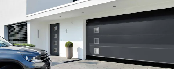 Strakke Hörmann garagedeur met vierkante glaspanelen bij moderne witte woning met zwarte auto op de oprit.