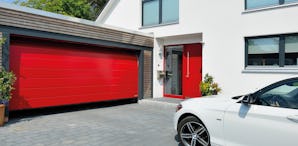 Med vår konfigurator får du snabbt och enkelt en överblick... klarröd entrédörr och garageport, på uppfarten står en bil.