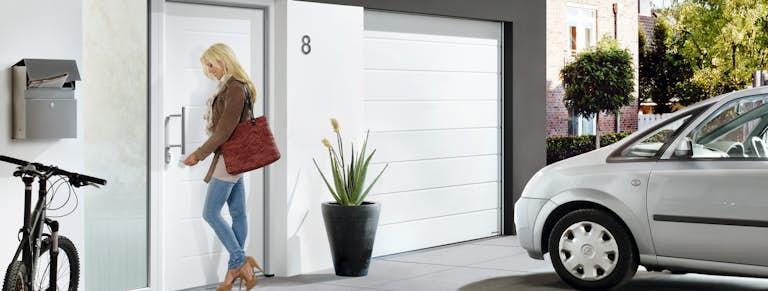 Dörrar - Entrédörrar Kvinna låser upp den vita entrédörren som matchar garageporten. På uppfarten står en bil och en cykel parkerad.