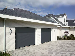 Takrullporten RollMatic OD sparar mer plats i taket än en... Mörk rullport till ett vitt tegelgarage bredvid står det stora boningshuset i vitt tegel.