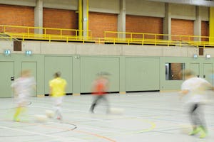 En sporthallsport ska stå emot mycket – från bollsparkar... ungdomar spelar fotboll i en idrottshall. Under läktaren på långsidan är gröna portar.